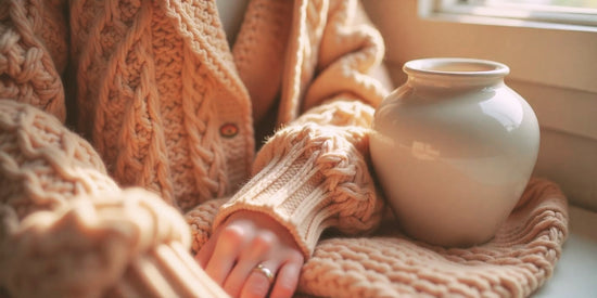 Close-up macro shot of a cozy knitted sweater with intricate patterns, featuring a warm color palette of browns and creams, with soft textures visible. An elegant, minimalistic urn made of smooth ceramic is placed beside the sweater.