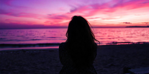 Young Middle-Eastern woman from behind with voluminous, dark hair, sitting on a bench made of driftwood, looking out at the sea, vibrant purple and pink sky reflecting on the water during sunset.